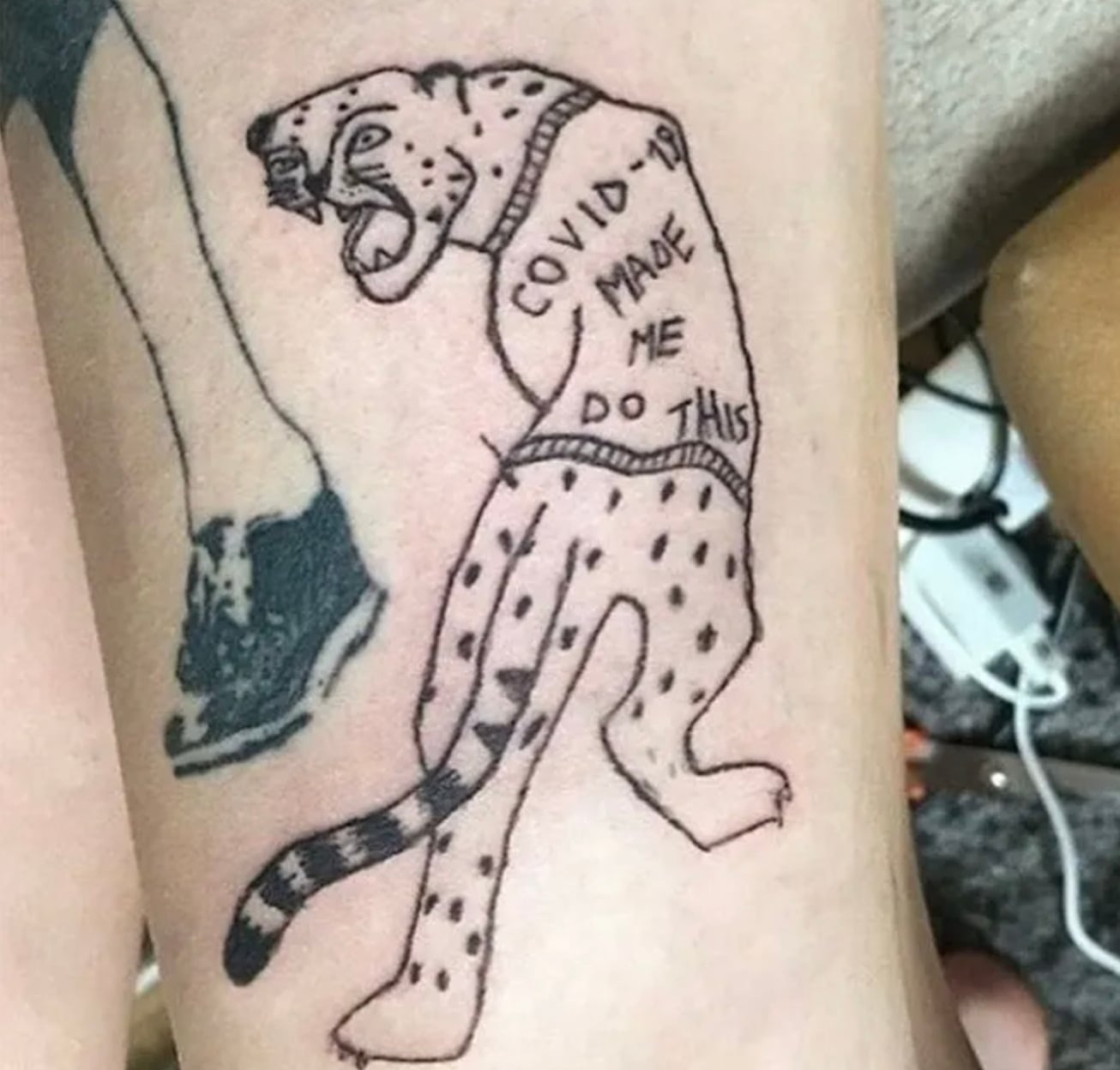 A tattoo of a leopard standing on two legs, snarling, with the words "COVID-19 MADE ME DO THIS" written on its body. There is also part of another tattoo showing a shoe and leg nearby.