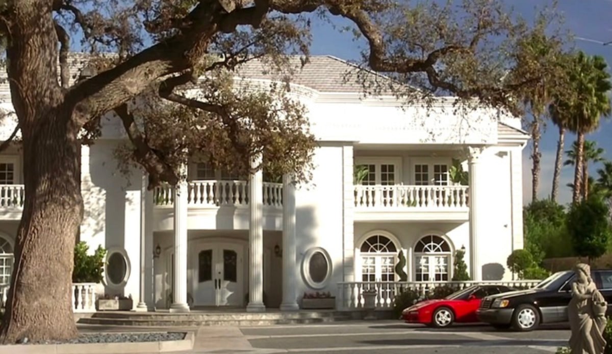A large white mansion with tall columns and balconies, partially shaded by a large tree, with a red sports car and a black sedan parked in front. The sky is clear and blue.
