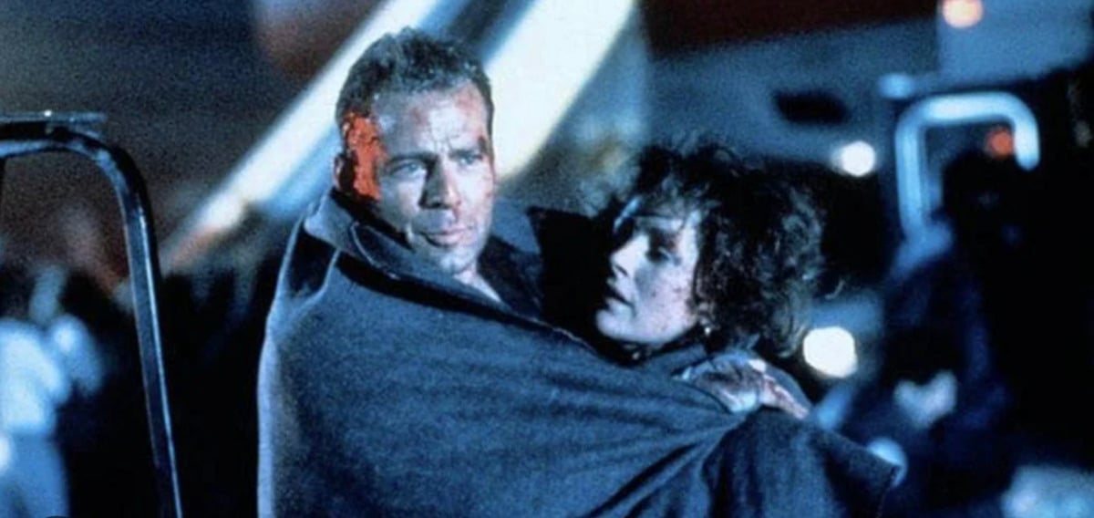 A man with a bruised face, wrapped in a gray blanket, carries an injured woman who is also covered in a blanket. The scene is at night, with emergency lights and vehicles blurred in the background.