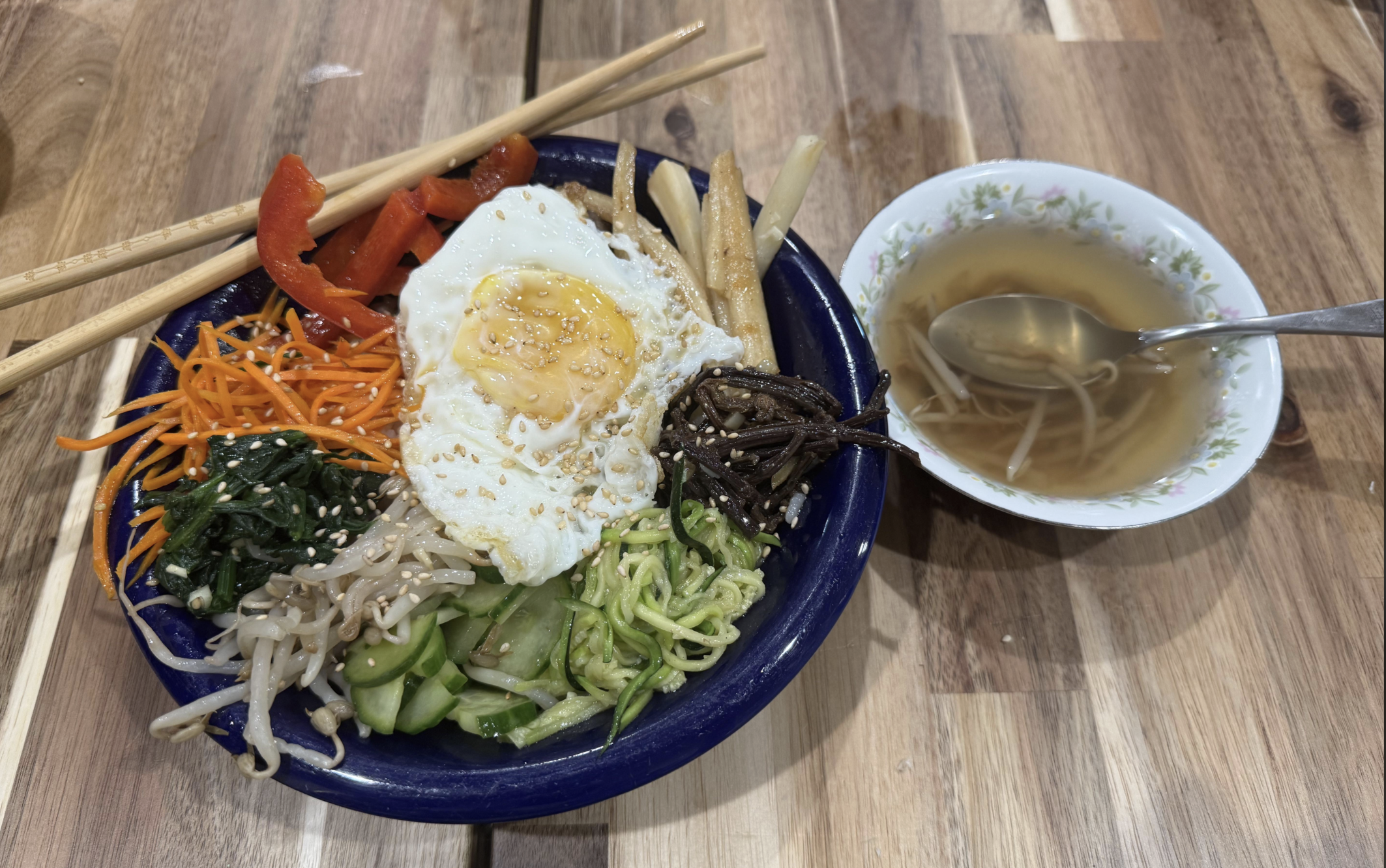 A colorful bibimbap bowl with shredded carrots, spinach, cucumber, bean sprouts, bell pepper, bamboo shoots, mushrooms, and a sunny-side-up egg, next to a small bowl of broth with bean sprouts and a spoon.