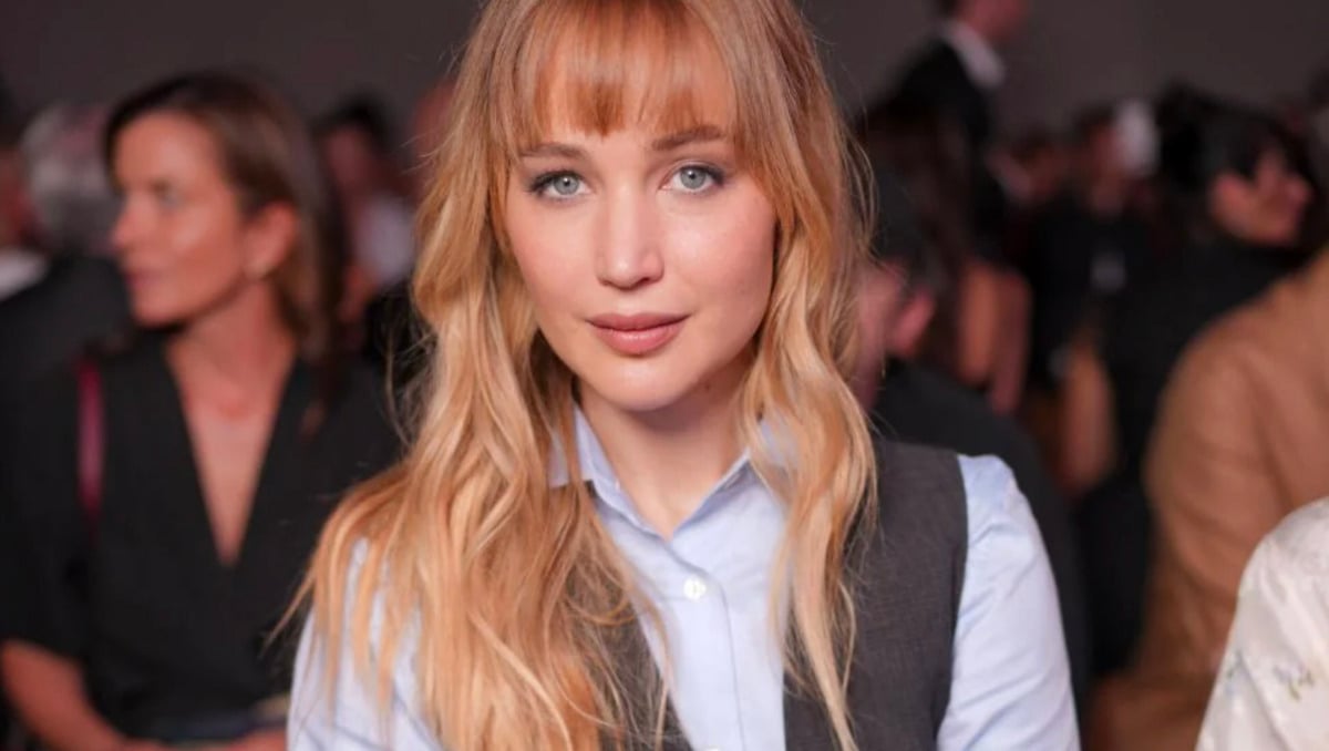 A woman with long blonde hair and bangs, wearing a light blue shirt and a dark vest, sits in a crowded room, looking directly at the camera. The background is softly blurred with people seated around her.