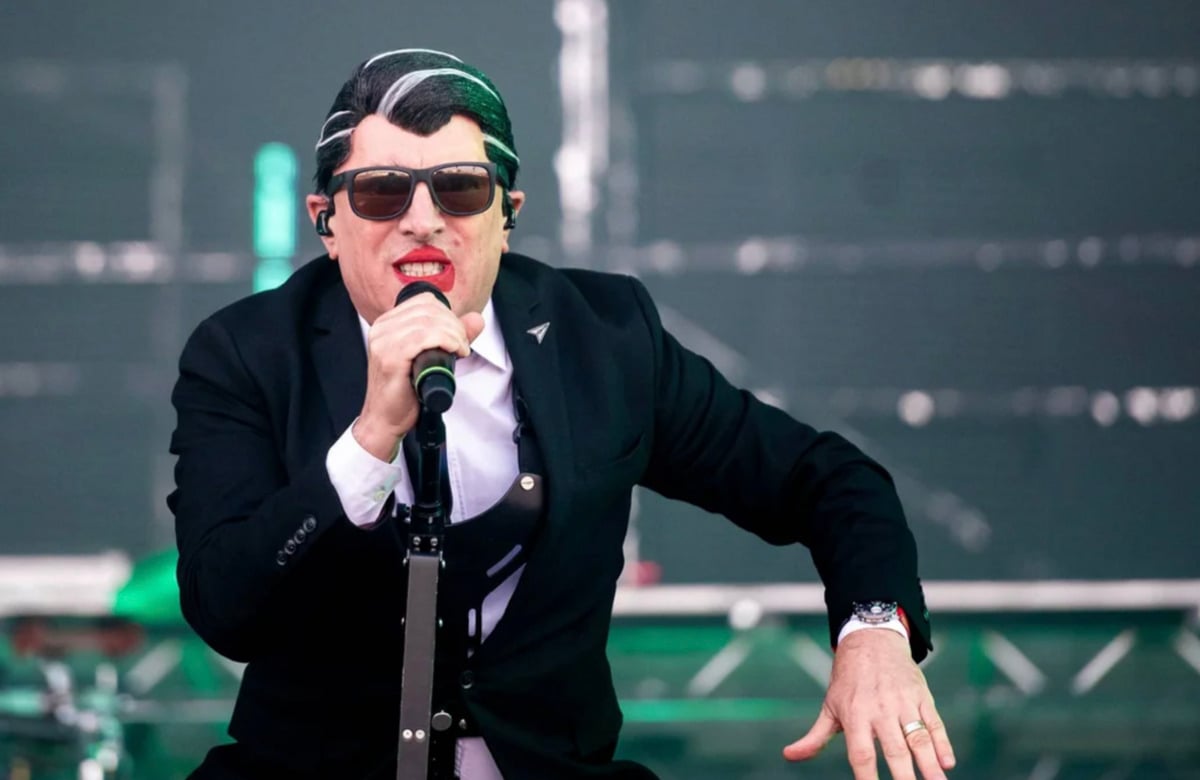 A person wearing a dark suit, sunglasses, and a mask with slicked-back hair and red lipstick holds a microphone while performing on stage, with a blurred background and green lighting.