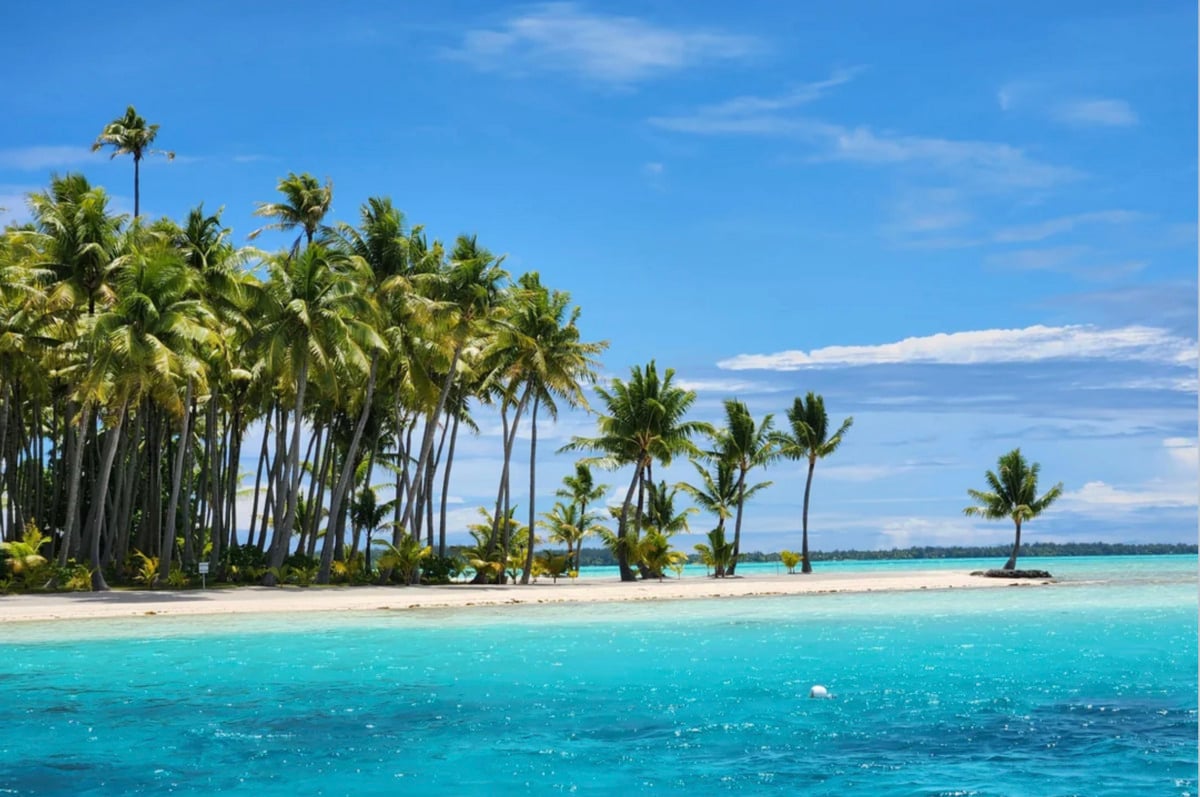 A tropical island with tall palm trees and white sandy beach surrounded by clear turquoise blue ocean under a bright sunny sky with scattered clouds.