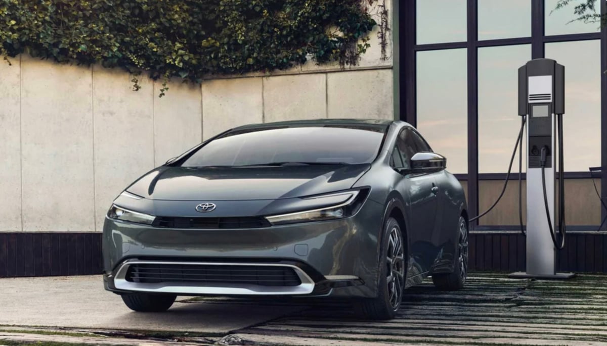 A sleek, dark gray Toyota sedan is parked next to an electric vehicle charging station outside a modern building with large windows and green vines on the wall.