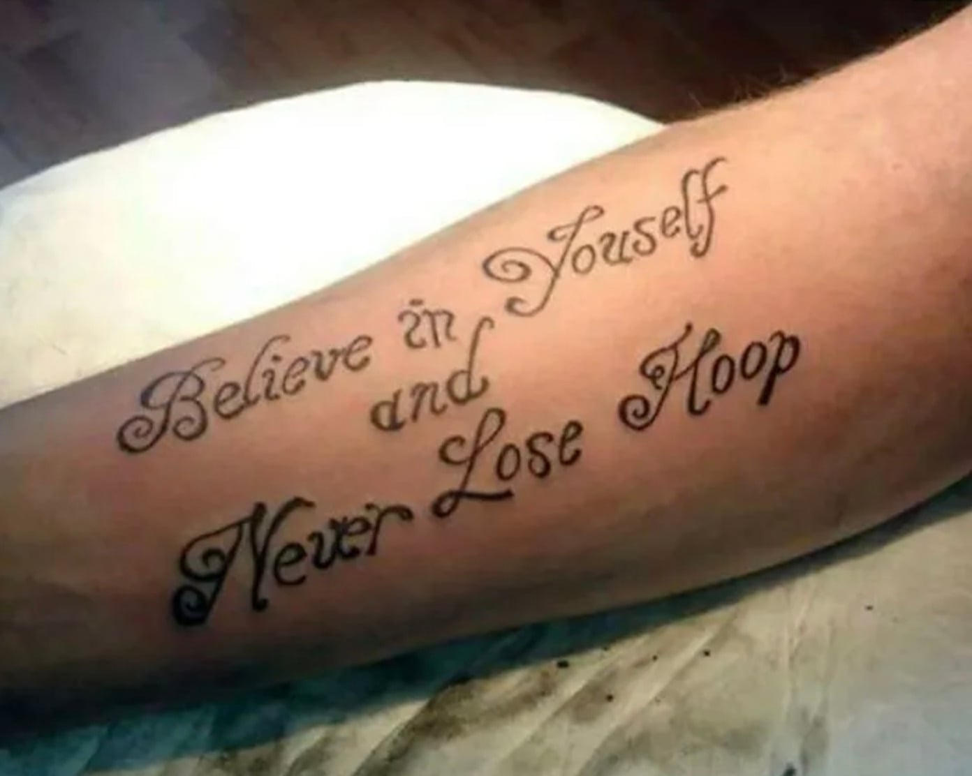 A tattoo on an arm reads, “Believe in Youself and Never Lose Hoop,” with “Youself” and “Hoop” misspelled instead of “Yourself” and “Hope.” The text is in decorative cursive font.