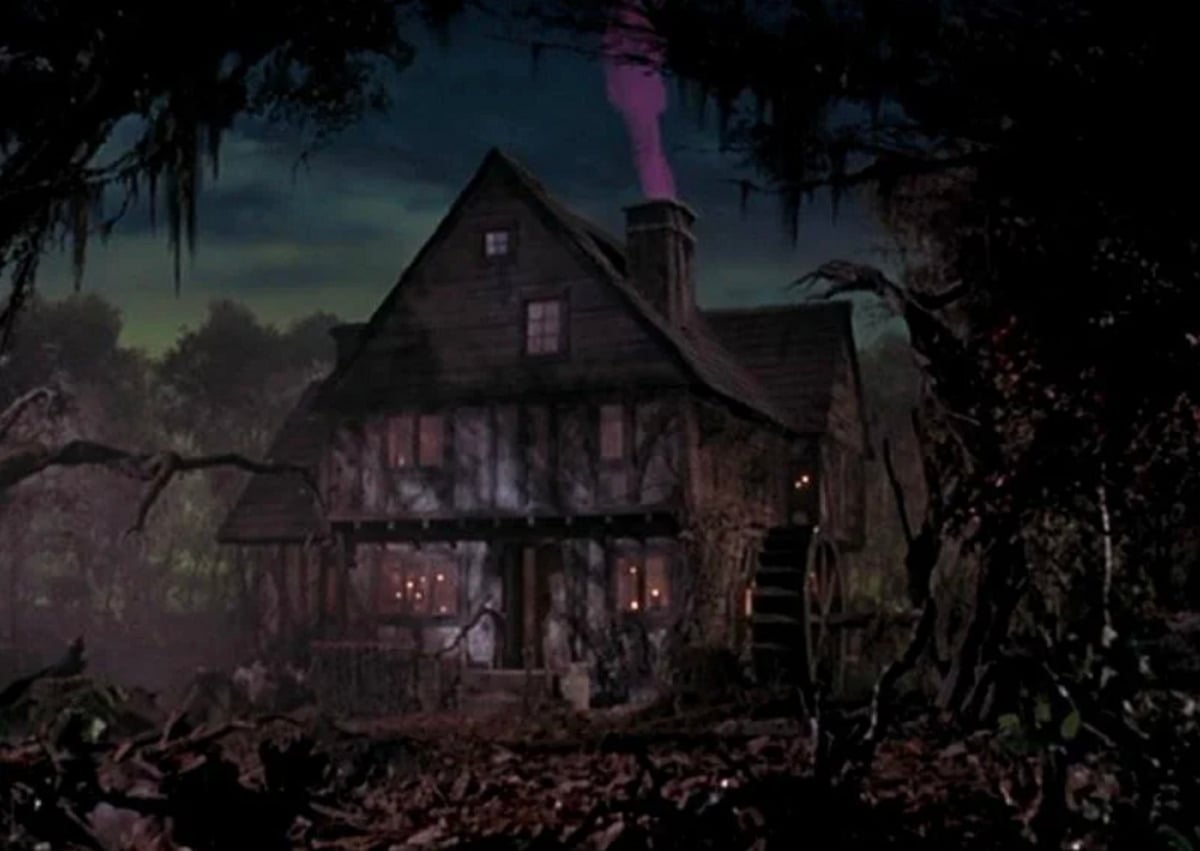 A dark, eerie house with a waterwheel sits in a foggy, swampy forest at night. Pink smoke rises from the chimney, and warm lights glow from the windows, creating a spooky atmosphere.