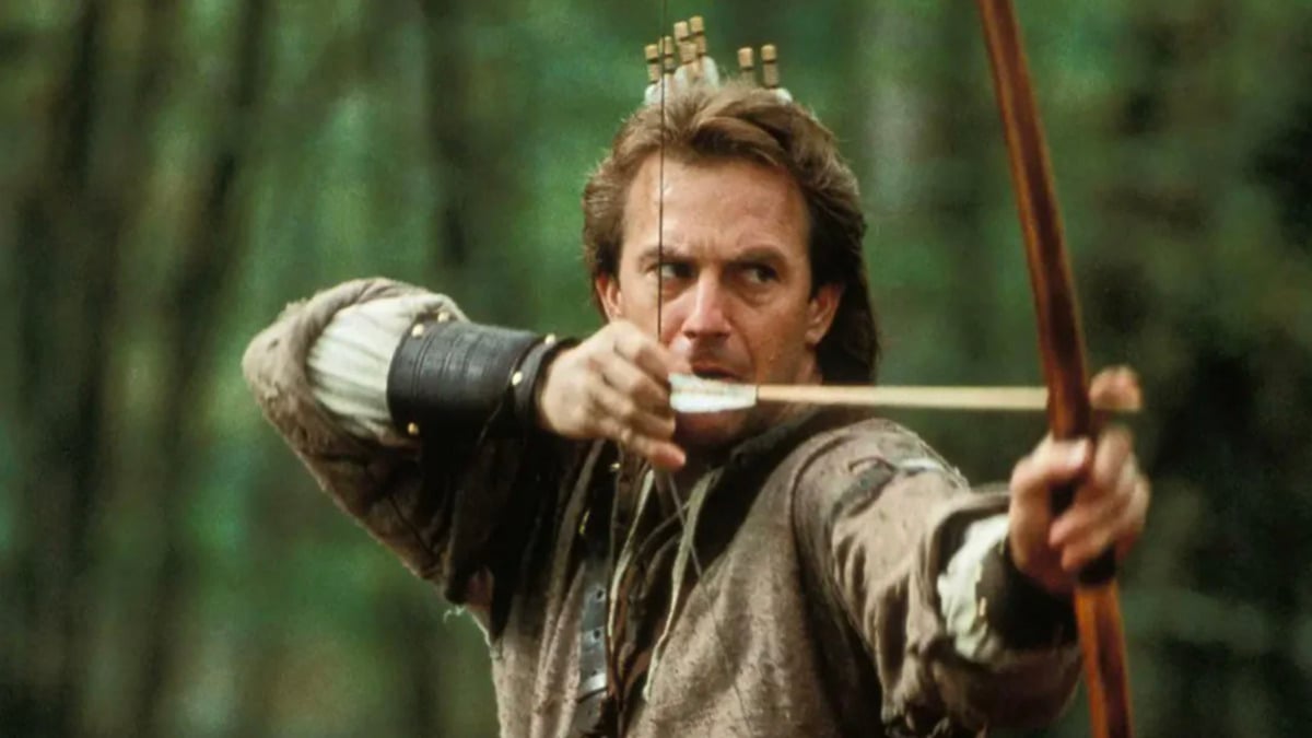 A man in medieval clothing draws a bow and aims an arrow intently, standing in a forest with arrows in a quiver on his back.