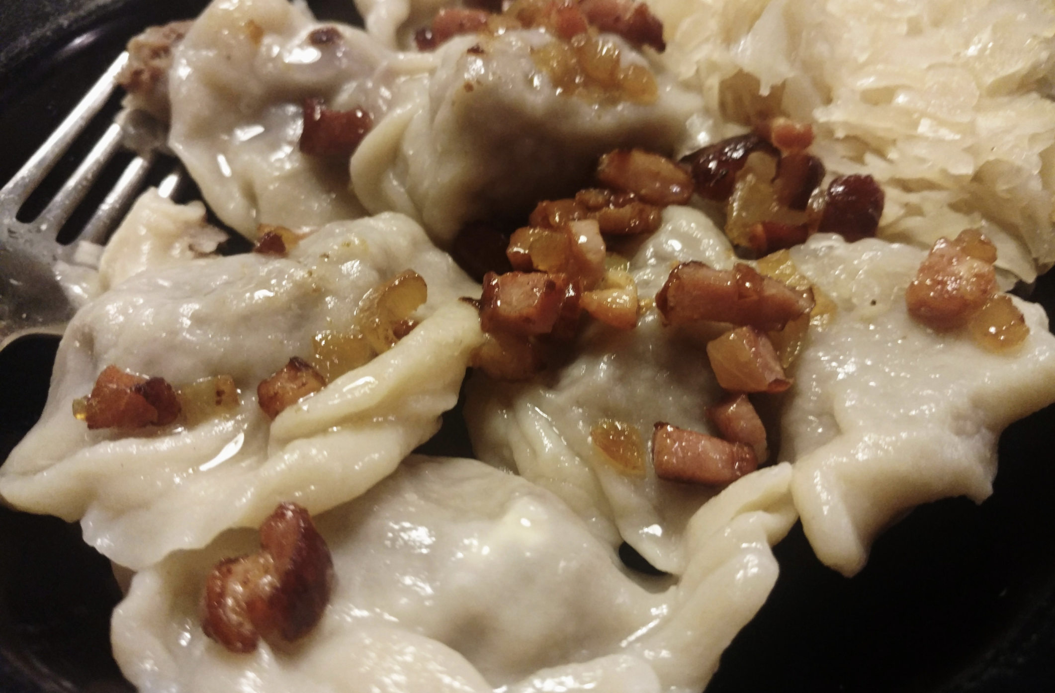 Close-up of pierogi topped with crispy diced bacon and sautéed onions, served alongside sauerkraut on a black plate. A fork is partially visible on the left side of the image.