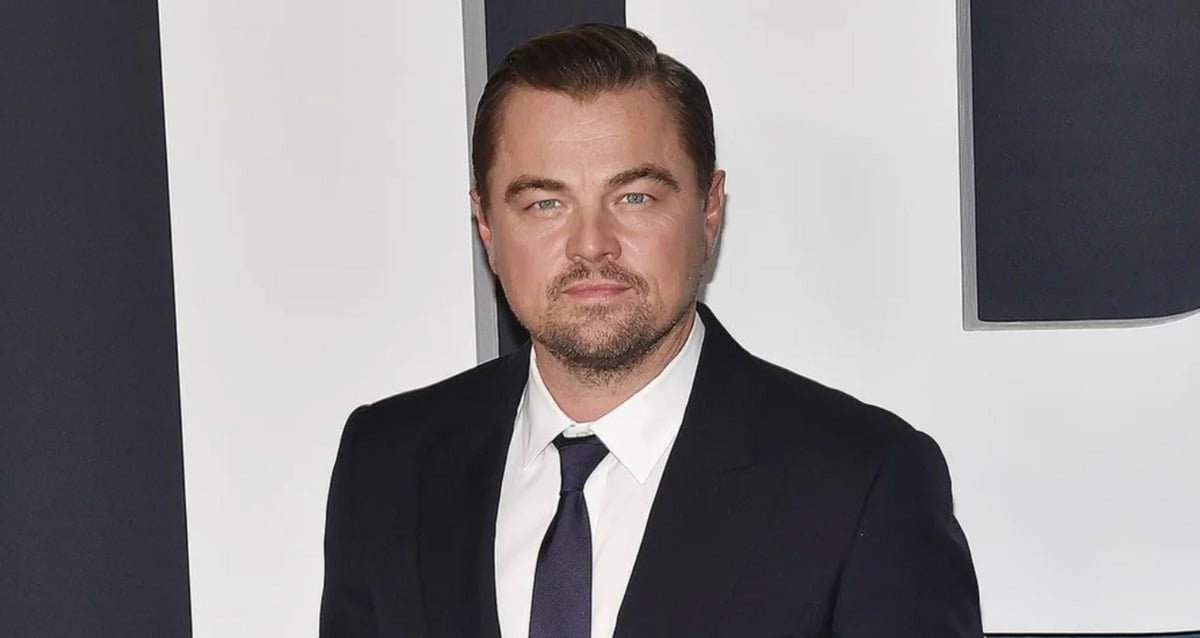 A man in a dark suit and tie stands in front of a black and white background, looking directly at the camera with a neutral expression.