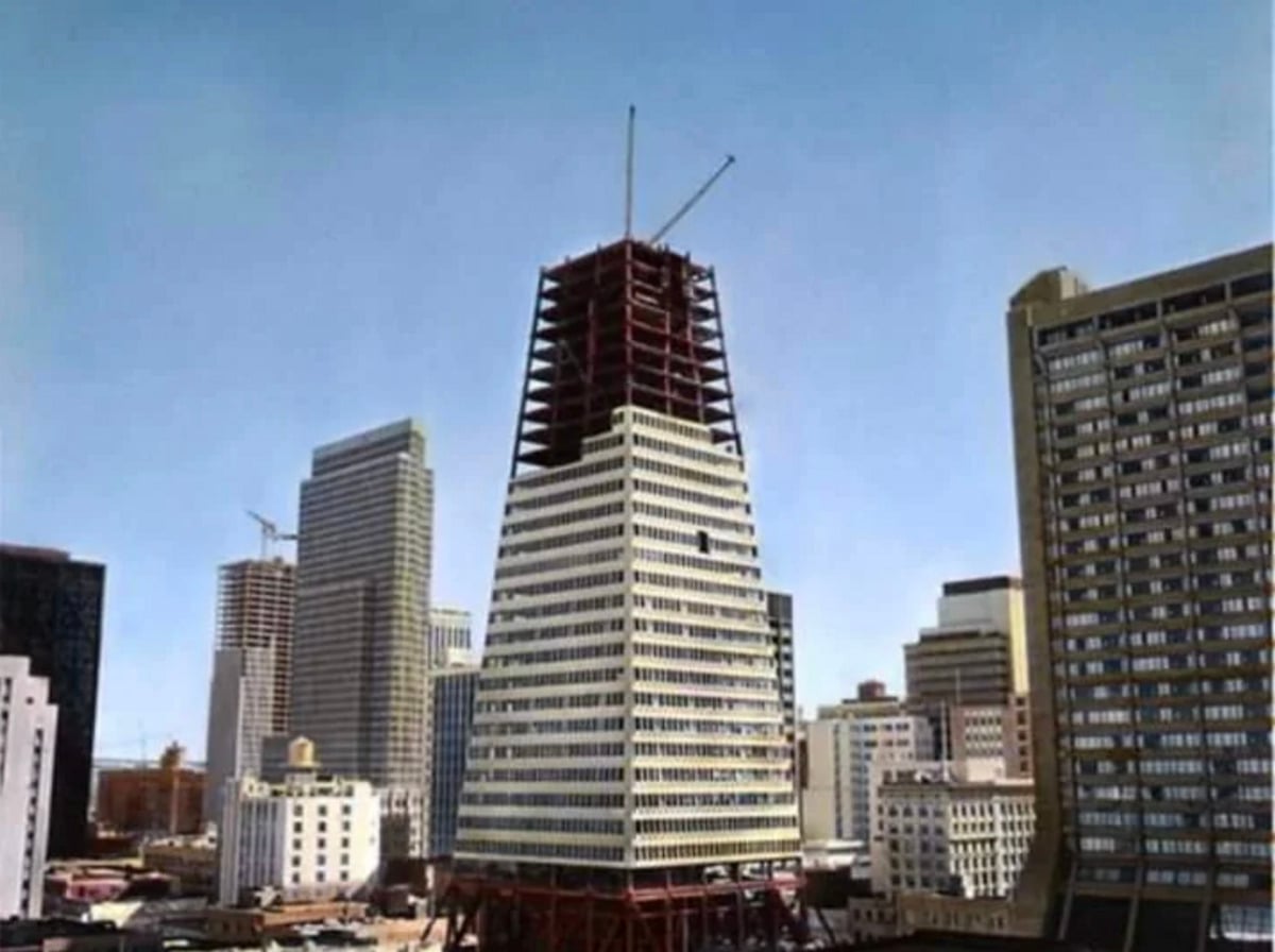 A tall skyscraper under construction with a unique tapered shape, surrounded by other completed high-rise buildings in a city skyline under a clear blue sky.
