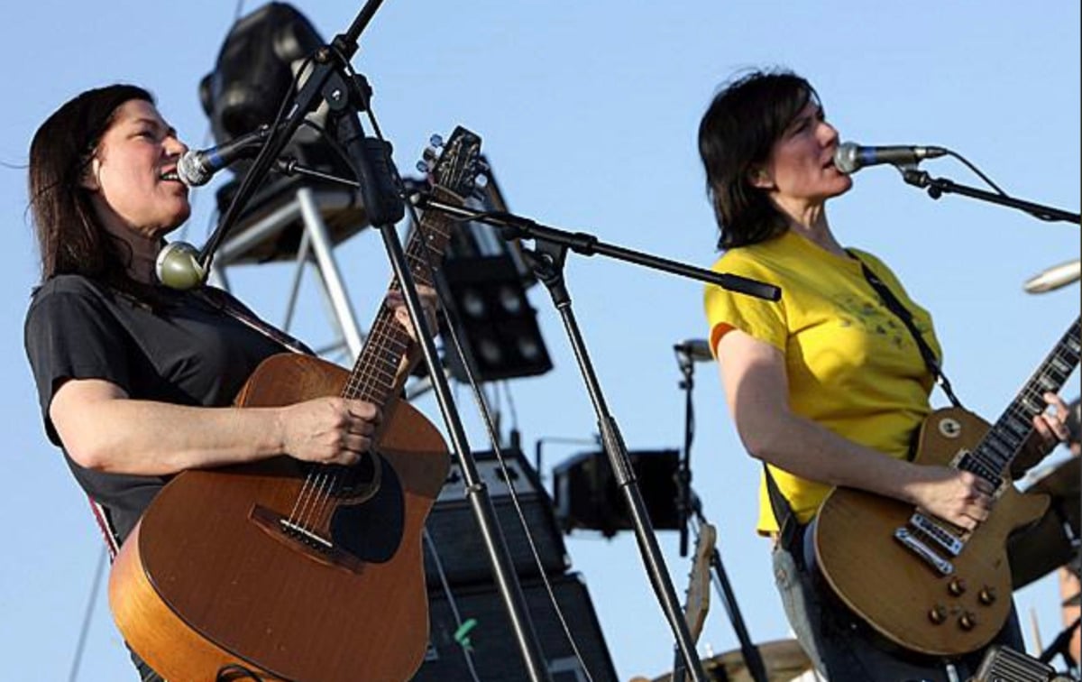 Two women play guitars and sing into microphones on an outdoor stage, with audio equipment and a blue sky in the background.