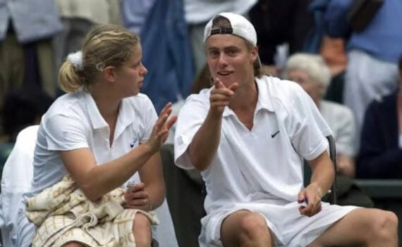 Two tennis players in white outfits sit on a bench, one gesturing while the other points and smiles. They appear to be having a conversation during a break, with spectators in the background.