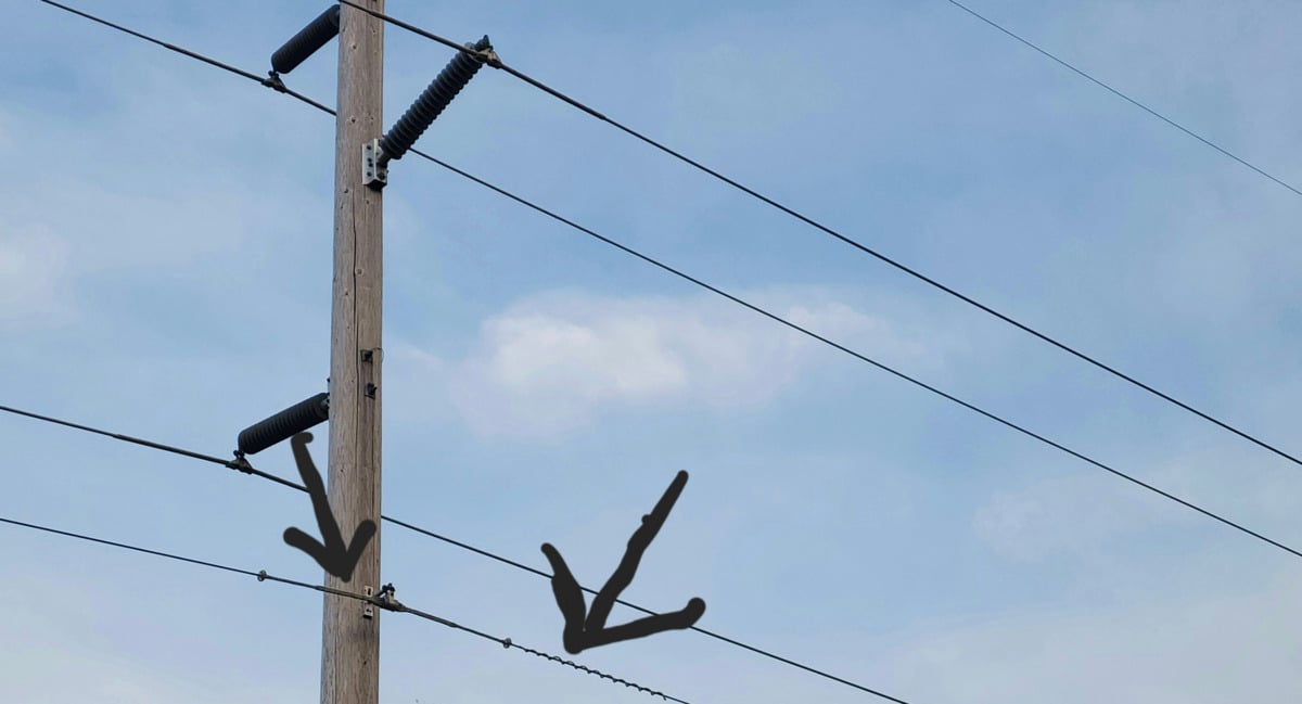 A wooden utility pole with three power lines against a blue sky. Two large black arrows point to two of the wires near the pole.
