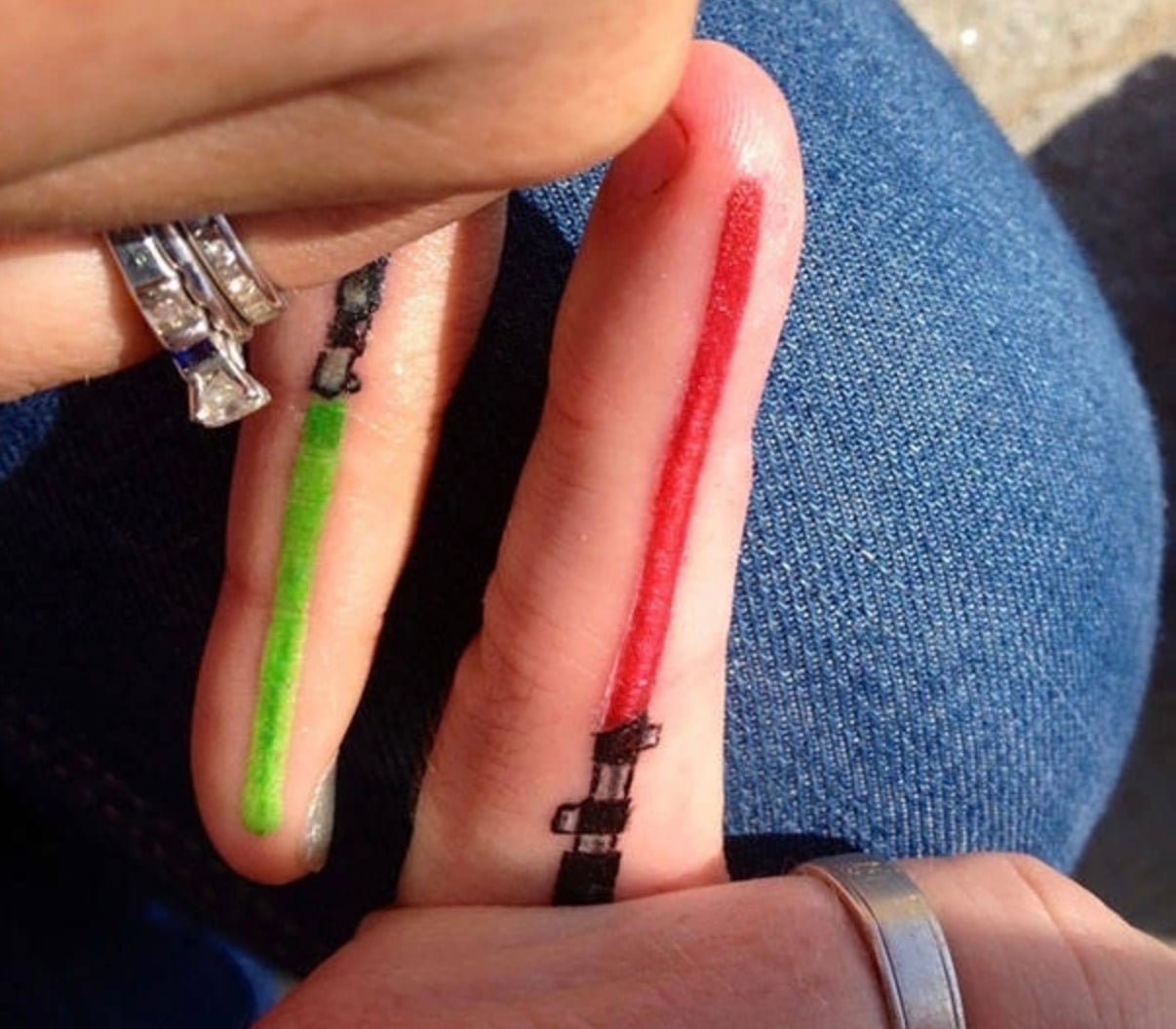 Two fingers with tattoos resembling lightsabers, one green and one red, are held side by side. The tattoos include black handles at the base. The hands wear silver rings and rest on blue denim fabric.