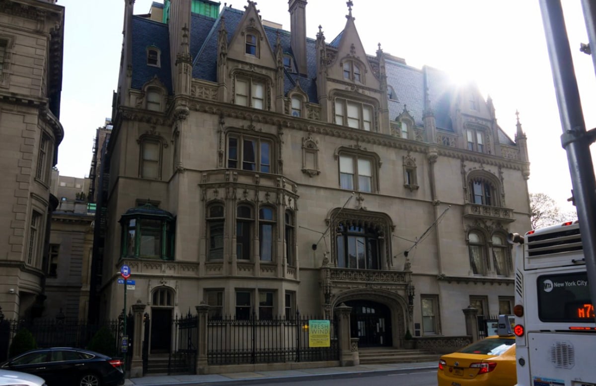 A large, ornate stone mansion with pointed roofs and detailed gothic-style architecture stands on a city street with a yellow taxi and a white bus passing by in front.