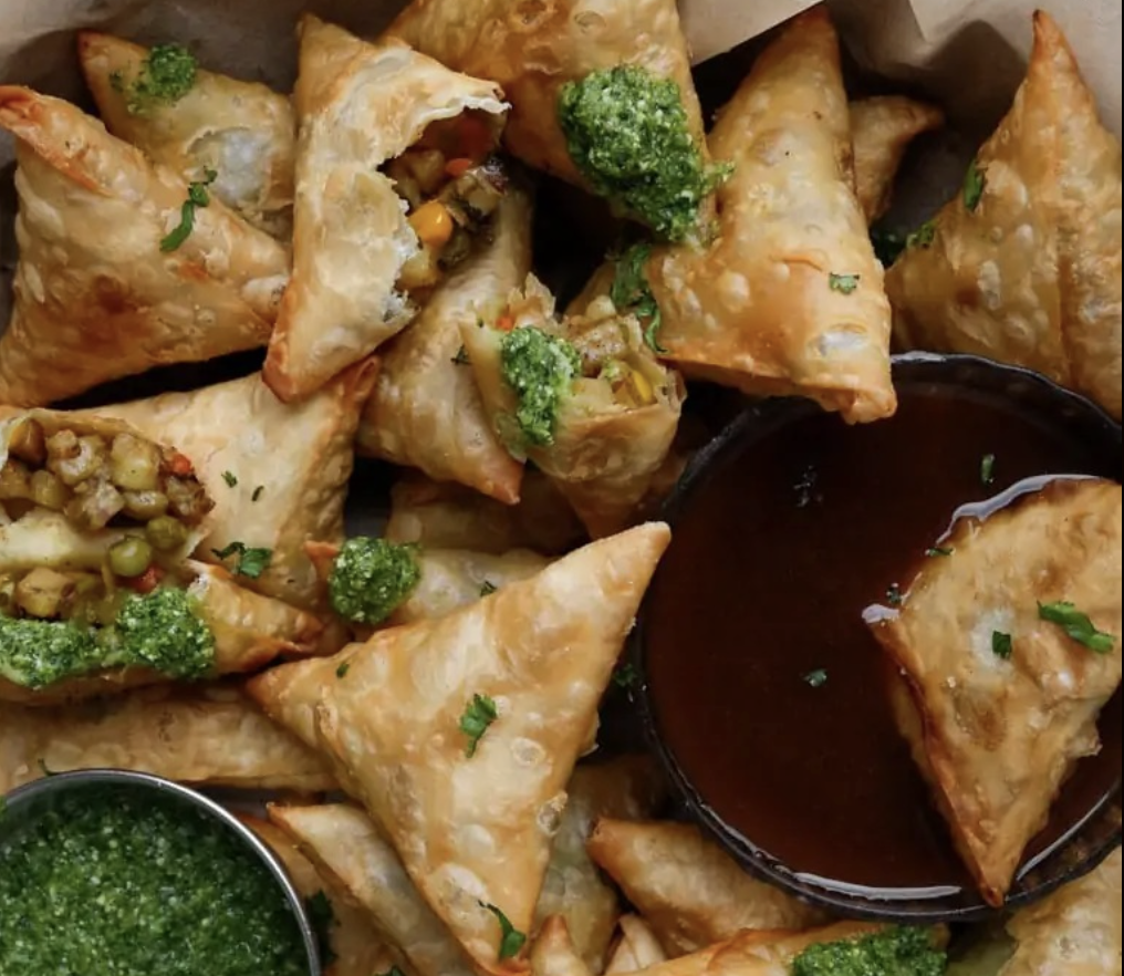 A pile of golden, crispy samosas filled with spiced vegetables, topped with green chutney, and served with a side of reddish-brown dipping sauce, arranged in a basket.