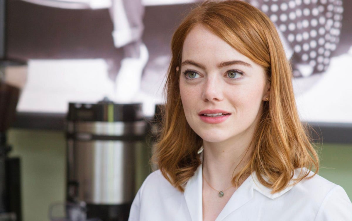 A woman with shoulder-length red hair, wearing a white shirt and a delicate necklace, stands indoors with a neutral expression. A coffee machine and a blurred black-and-white photo are visible in the background.
