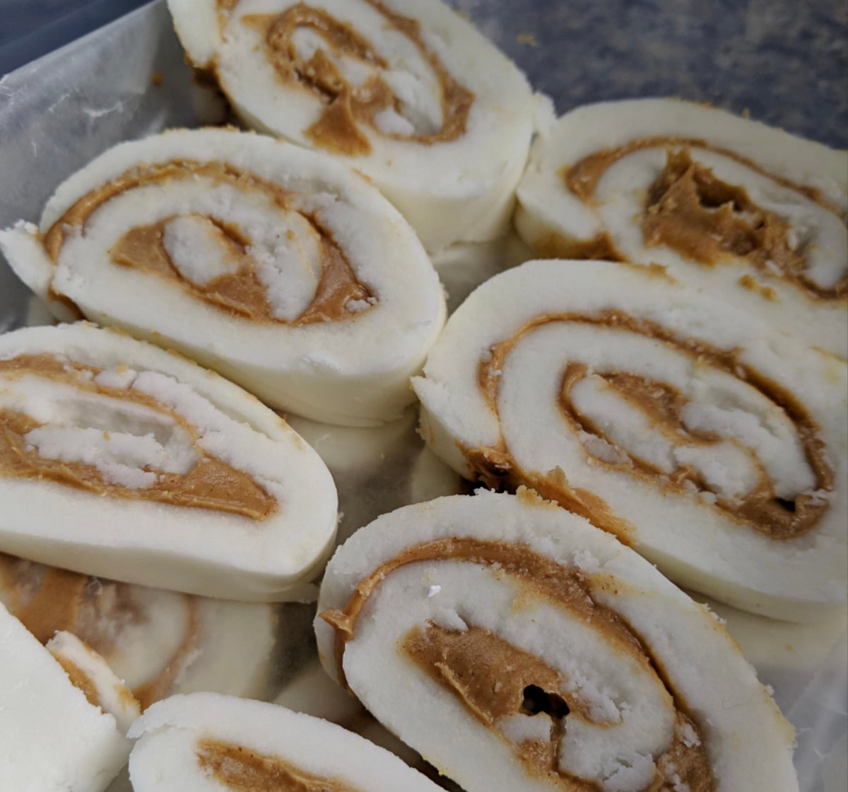 Close-up of several slices of peanut butter pinwheel candy, showing creamy peanut butter swirled inside white, rolled sugar dough. The candies are arranged closely together on a tray.