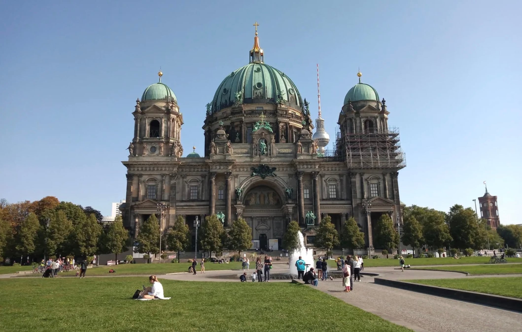 People relax and walk on a grassy lawn in front of a large historic cathedral with green domes and ornate architecture, under a clear blue sky. Some scaffolding is visible on the right dome.