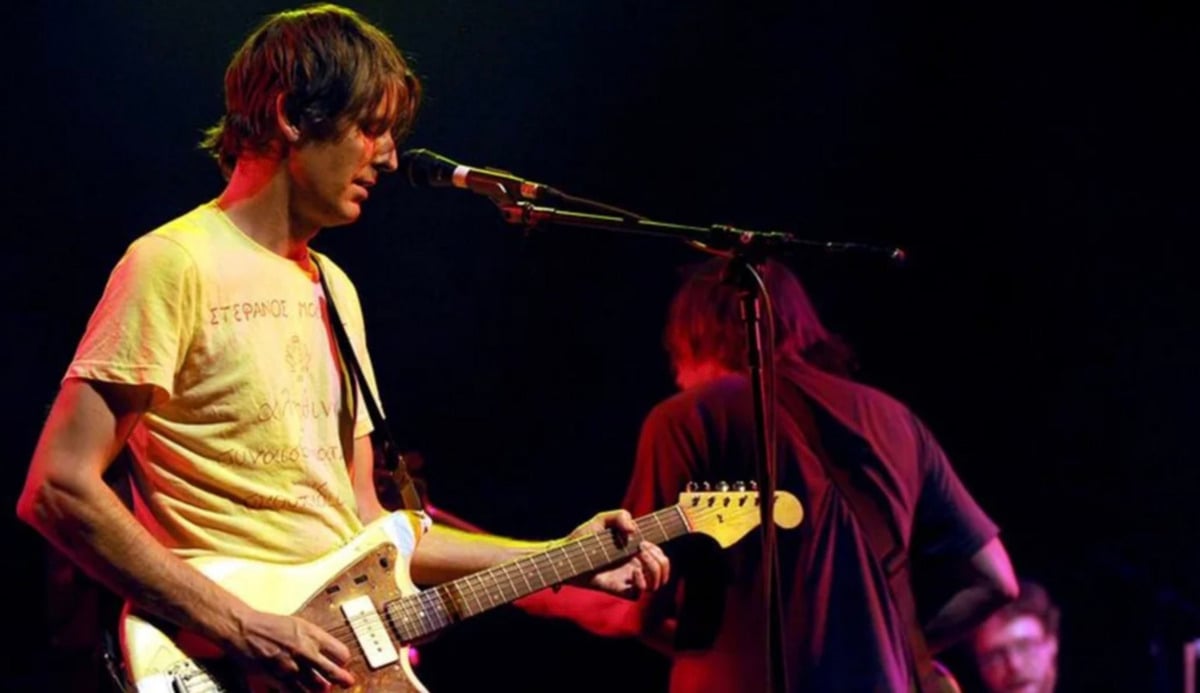 A musician wearing a yellow t-shirt plays an electric guitar and sings into a microphone on a dimly lit stage, while another guitarist stands slightly behind him, facing away.