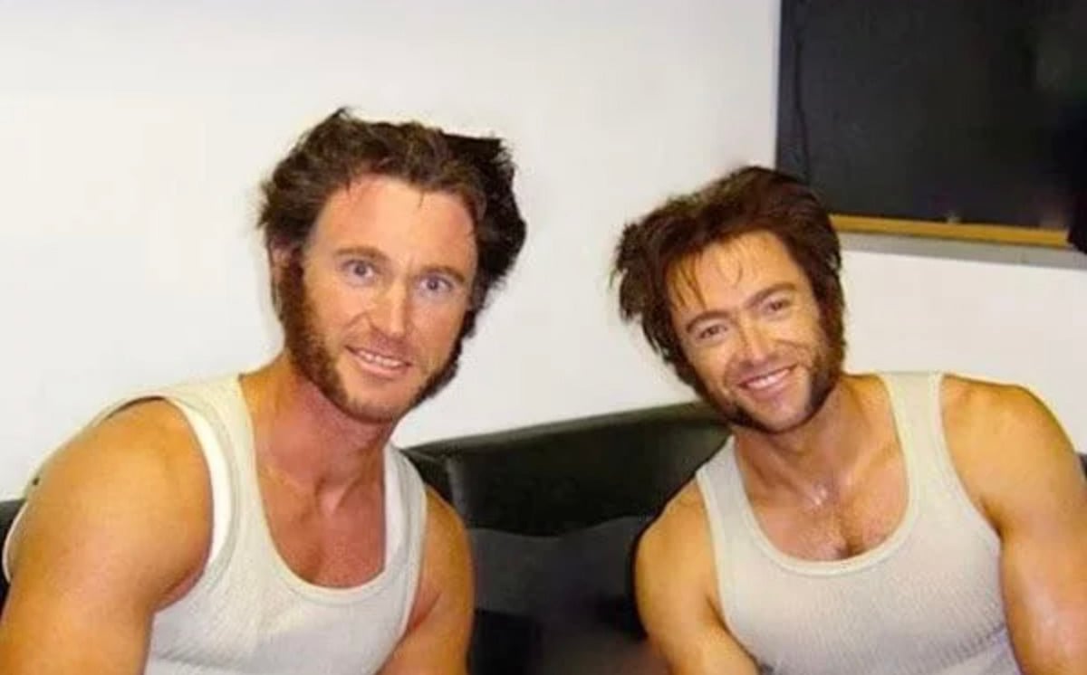 Two men with muscular builds, dressed in white tank tops and styled with thick, mutton-chop sideburns and tousled hair, sit side by side smiling in a casual indoor setting.