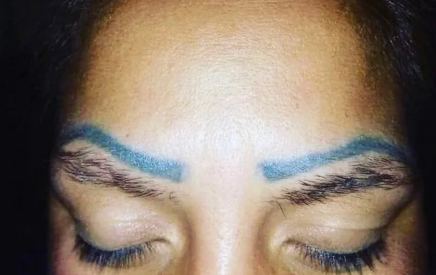 A close-up of a person's closed eyes and forehead, showing uneven blue eyebrow tattoos above their natural brows. The natural eyebrows are partially visible beneath the bold, bright blue tattoo lines.