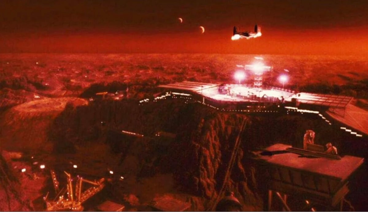 A futuristic colony under a red, alien sky features a central illuminated structure, distant flying vehicle, rocky terrain, and two moons visible above the horizon. The scene has a dramatic, otherworldly atmosphere.