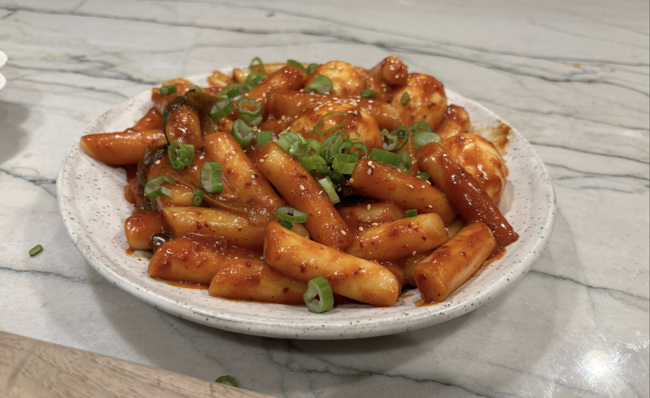 A plate of spicy Korean tteokbokki, featuring chewy rice cakes and boiled eggs coated in red sauce, garnished with chopped green onions, sits on a marble countertop.