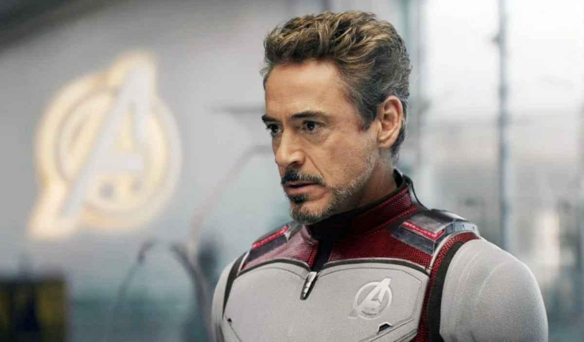 A man with short brown hair and a trimmed beard wears a high-tech, red and gray suit with an Avengers logo behind him, standing in a futuristic setting and looking serious.