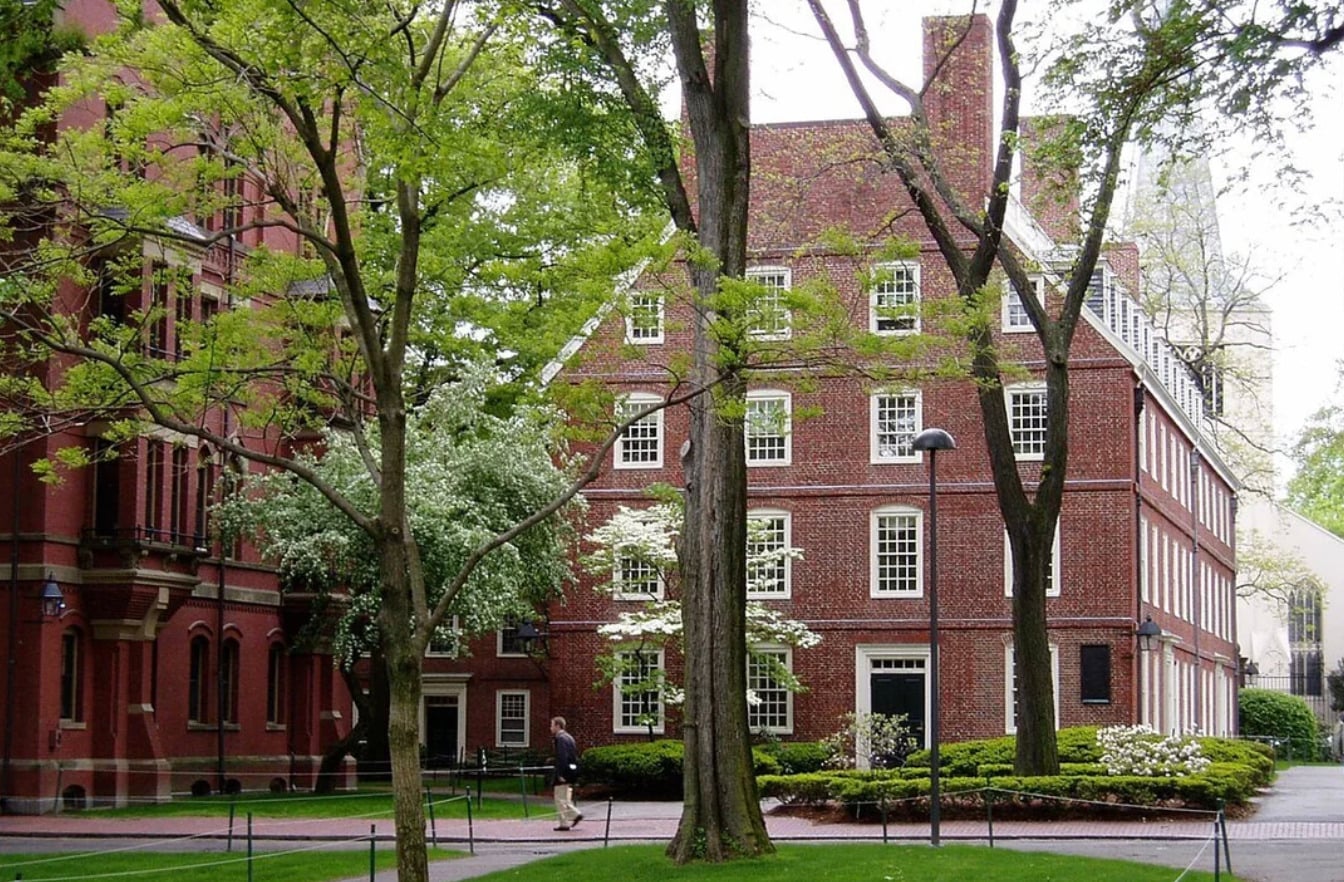 A person walks on a paved path beside a historic red brick building with white trim, surrounded by green trees and shrubs on a leafy college campus.