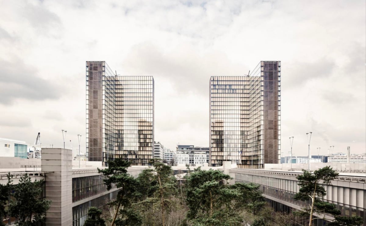 Two modern glass high-rise buildings face each other with a courtyard of trees and greenery in between, set against a cloudy sky and surrounded by urban structures.