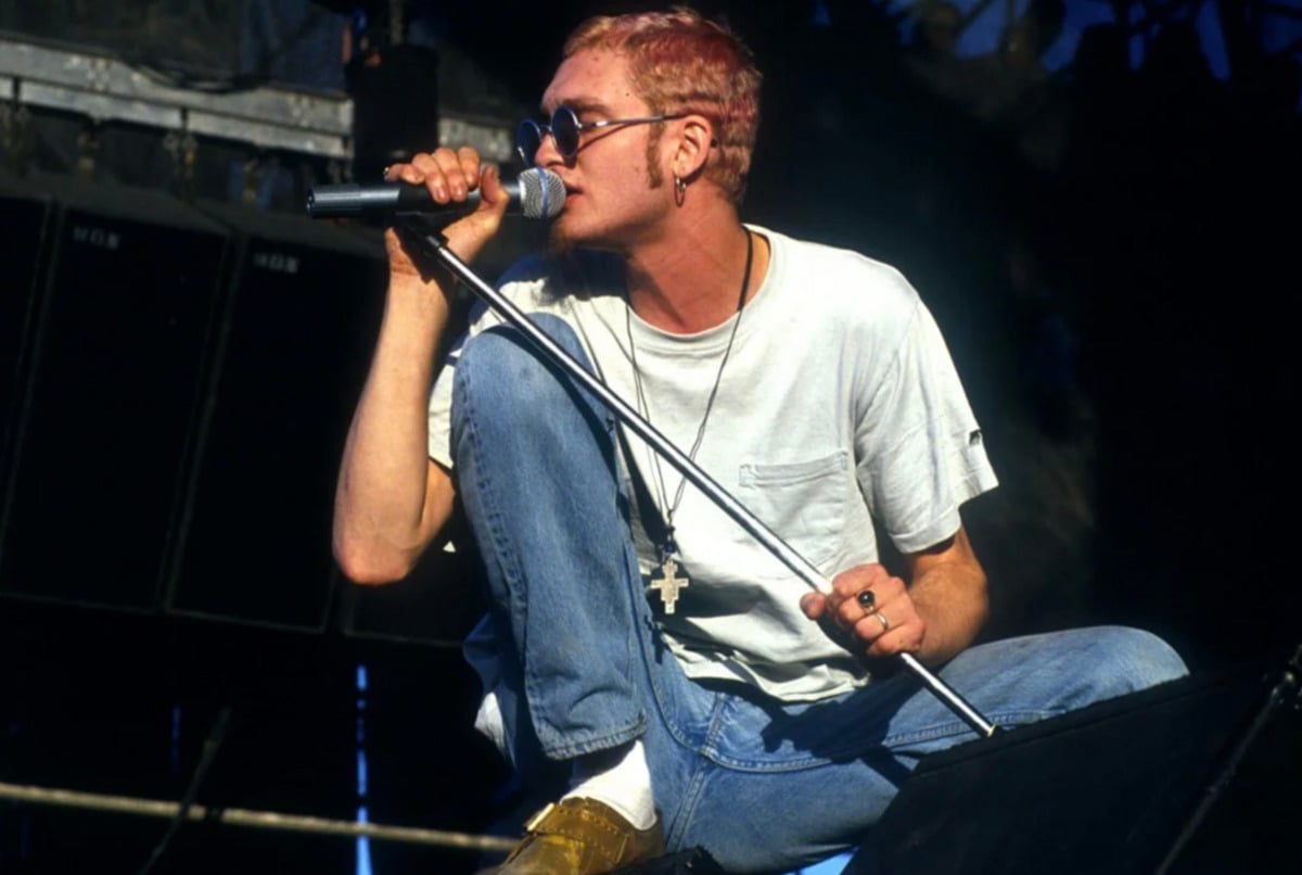 A musician with short, light hair and sunglasses crouches on stage, singing into a microphone. He wears a white t-shirt, blue jeans, and a necklace with a cross pendant, performing in front of stage equipment.