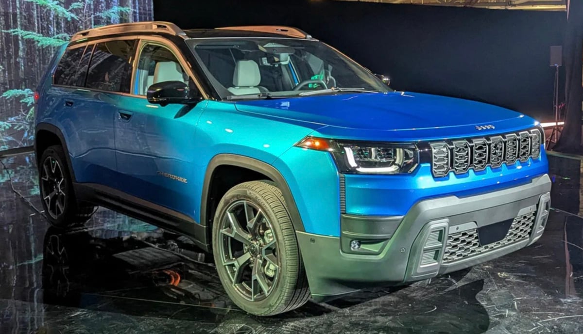 A blue Jeep SUV is displayed under bright lights on a glossy black floor, showcasing its modern design, black wheels, and signature grille. The vehicle is viewed from the front left angle.