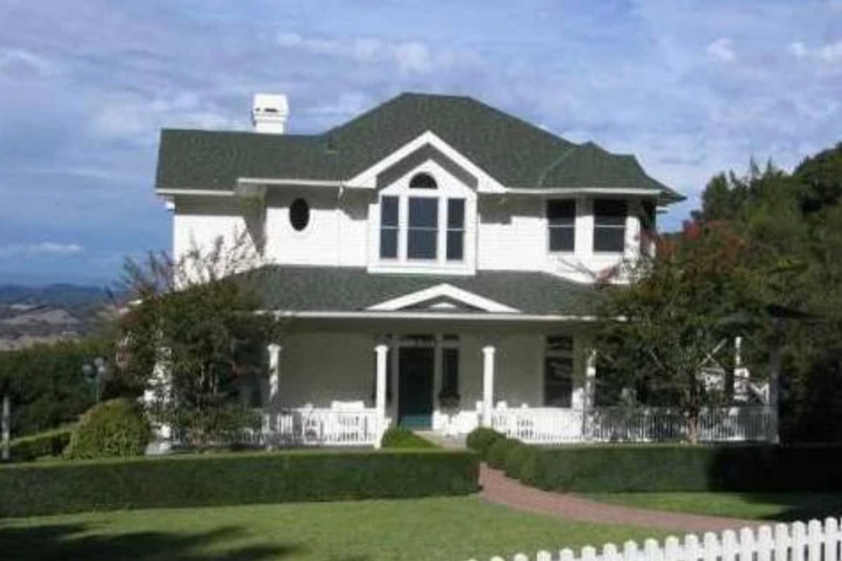A two-story white house with green roofing sits behind a white picket fence, surrounded by trimmed bushes and trees, with a manicured lawn and brick walkway leading to the front porch.