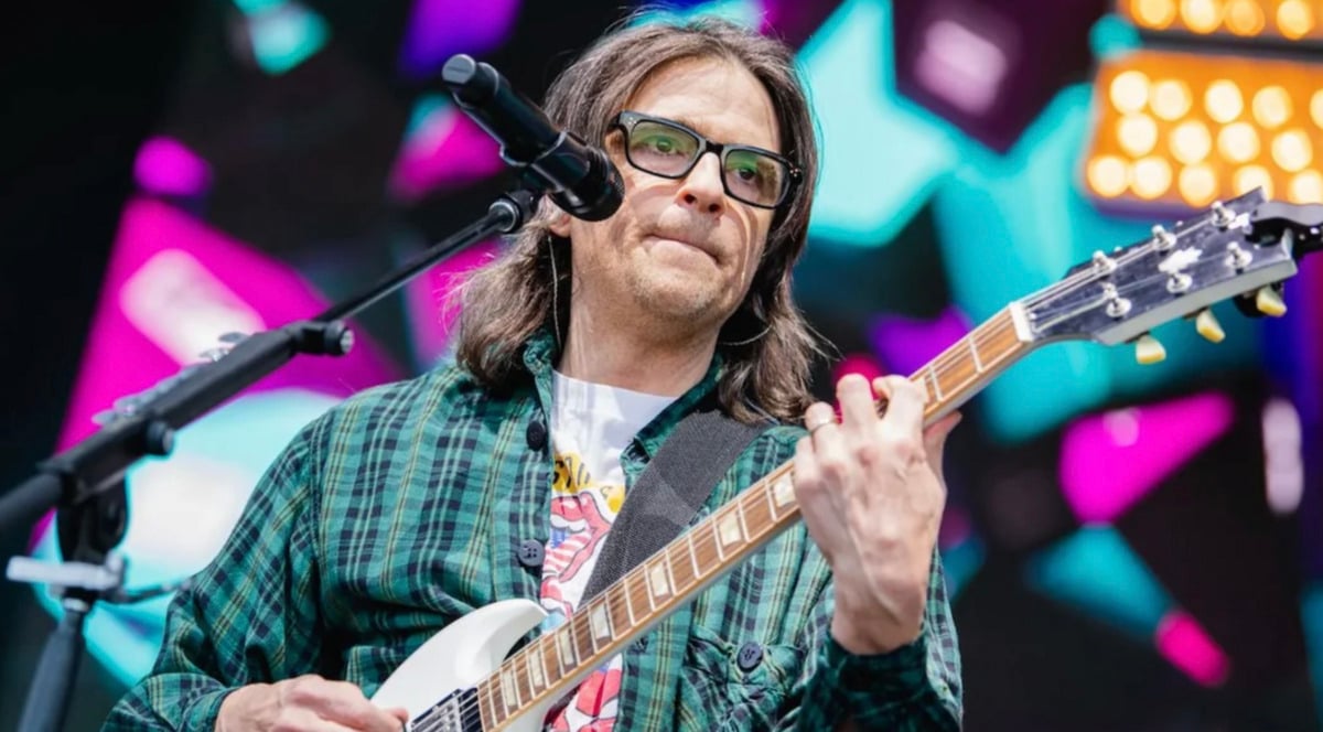 A musician with long brown hair and glasses plays an electric guitar and sings into a microphone on stage, with colorful geometric lights in the background.