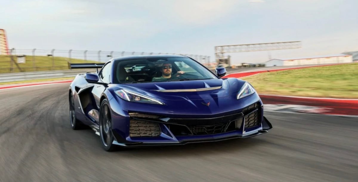 A blue sports car speeds around a racetrack, driven by a person wearing sunglasses. The car has sharp, angular features and bright headlights, with blurred scenery in the background indicating motion.