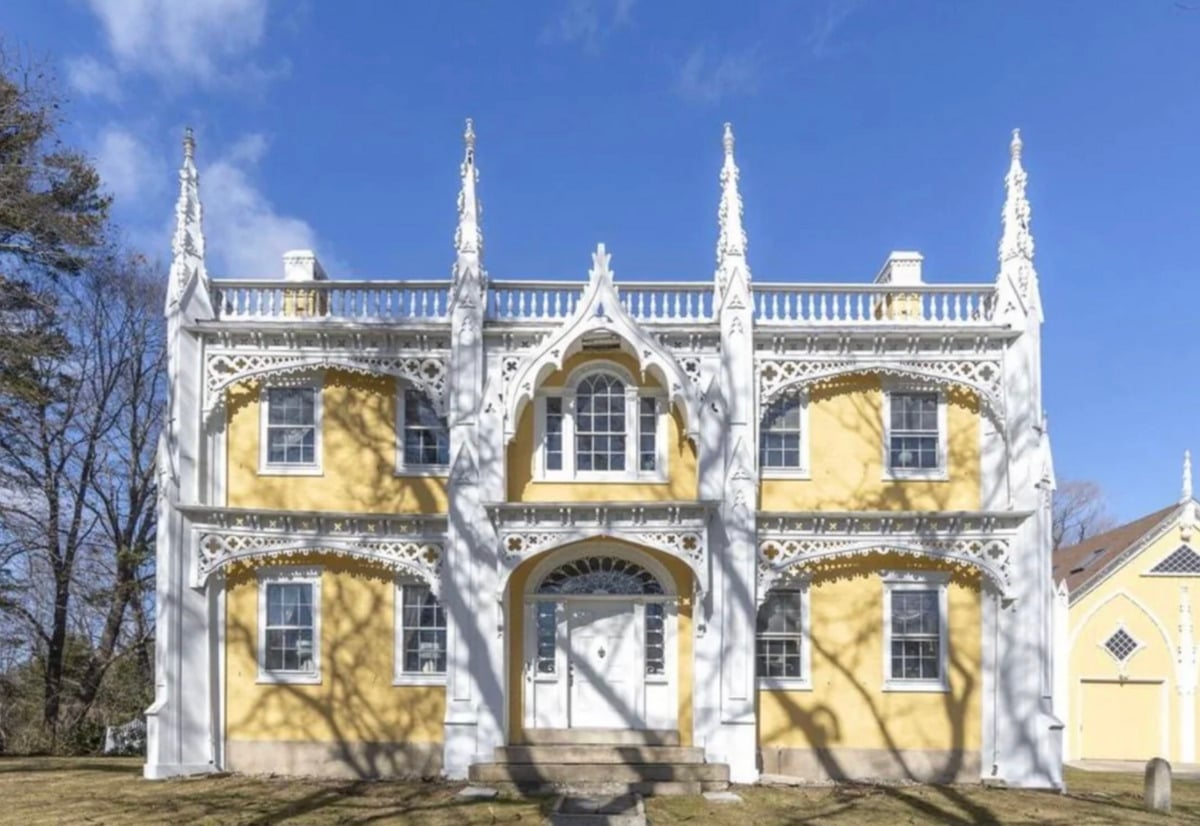 A yellow two-story house with white elaborate Gothic trim, pointed arches, and decorative spires, set against a blue sky with tree shadows cast on the front.