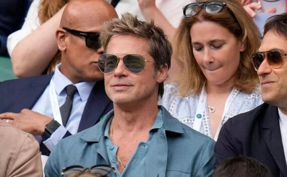A man wearing sunglasses and a blue shirt sits in a crowd at an outdoor event, surrounded by other people, some also wearing sunglasses and lanyards.