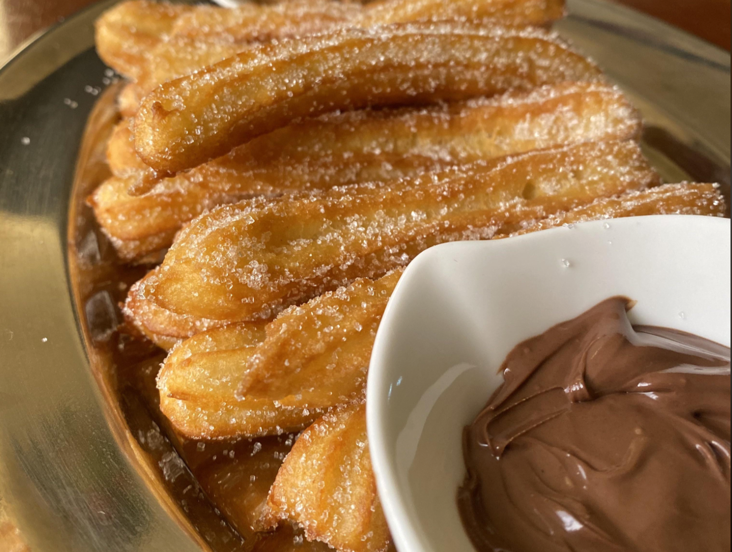 A plate of sugar-coated churros is stacked on a gold tray, served with a small white bowl filled with creamy chocolate dip.