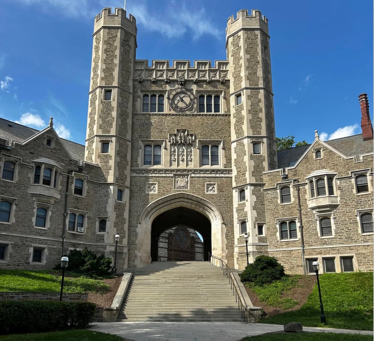 A large stone building with twin towers and a central arched gateway stands beneath a blue sky. The facade features a clock, intricate details, and wide steps leading up to the entrance.