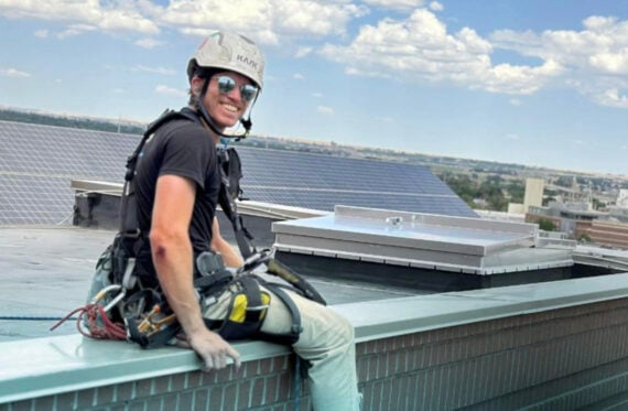 A person wearing safety gear and a helmet sits on the edge of a rooftop, smiling at the camera. The background shows solar panels, buildings, and a blue sky with scattered clouds.
