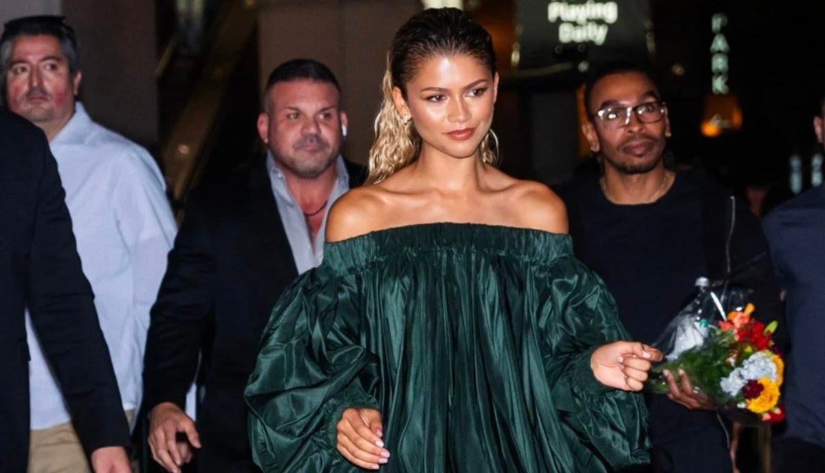 A woman in an off-the-shoulder, dark green, voluminous dress walks outside at night, surrounded by several people. One person is holding a bouquet of flowers. The woman has slicked-back hair and a confident expression.