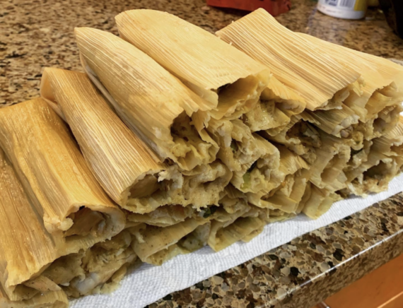 A stack of homemade tamales wrapped in corn husks rests on a paper towel atop a granite countertop. The tamales are golden and look freshly steamed, with some filling visible at the ends.