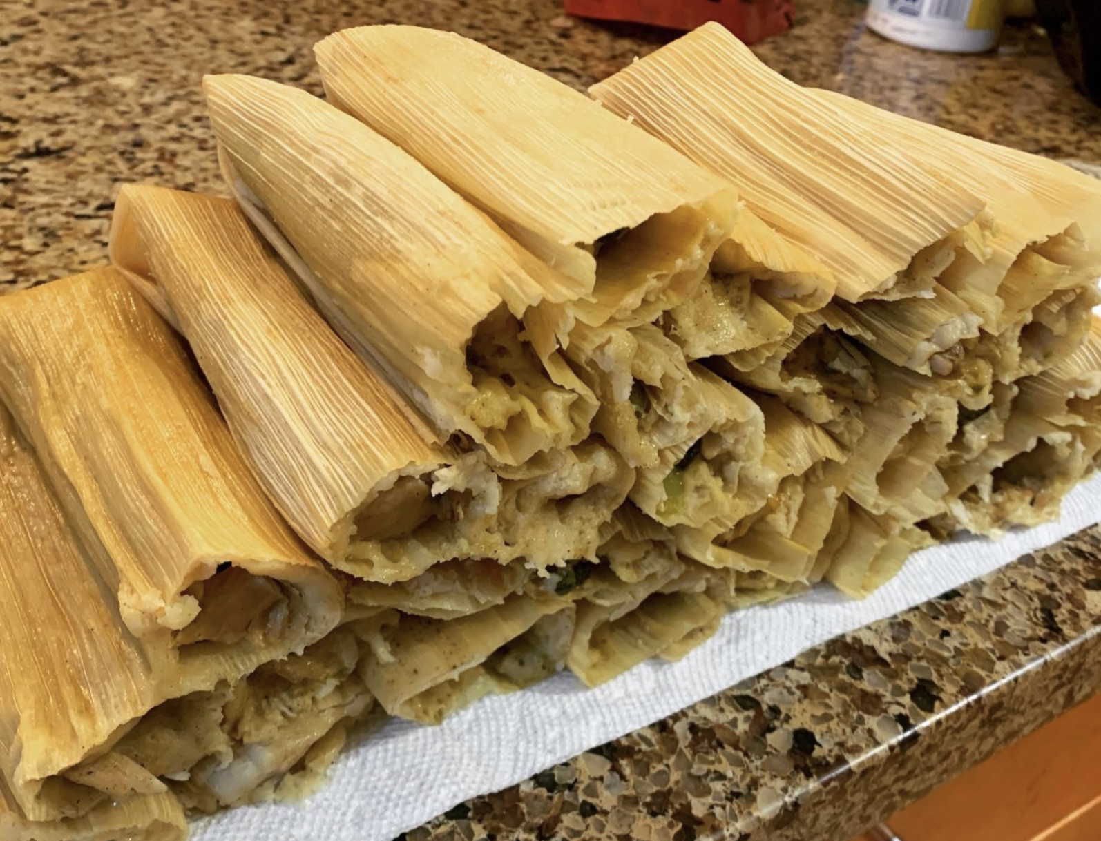 A stack of homemade tamales wrapped in corn husks rests on a paper towel atop a granite countertop. The tamales are golden and look freshly steamed, with some filling visible at the ends.