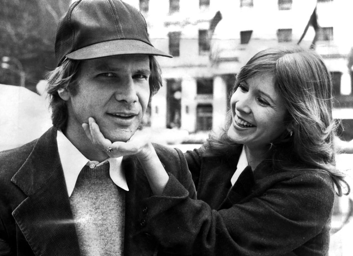 A woman smiles and gently holds a man's chin with one hand. The man wears a cap and a jacket, looking at the camera with a neutral expression. They are outdoors with buildings in the background.