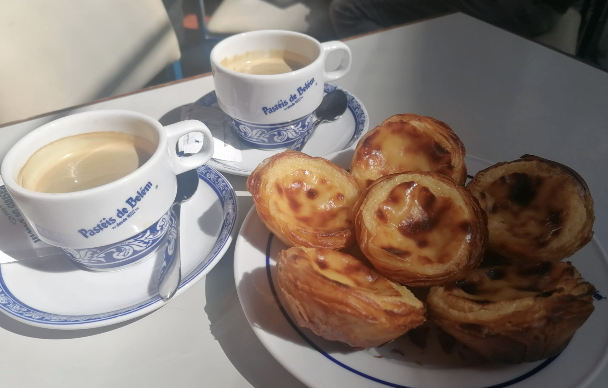 Two cups of coffee and a plate with several golden Portuguese custard tarts (pastéis de nata) on a white table, with sunlight casting shadows across the scene.