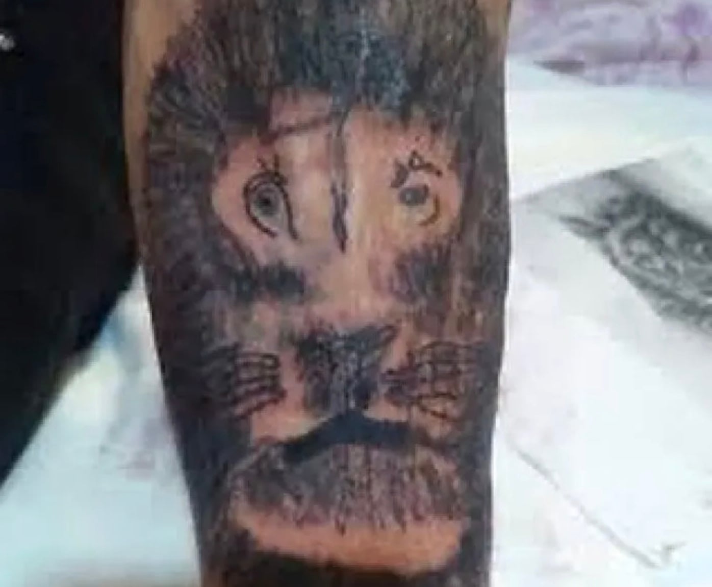 A tattoo of a lion’s face on a forearm, with uneven eyes, a lopsided nose, and an asymmetrical mane, giving the lion a distorted, cartoon-like appearance.
