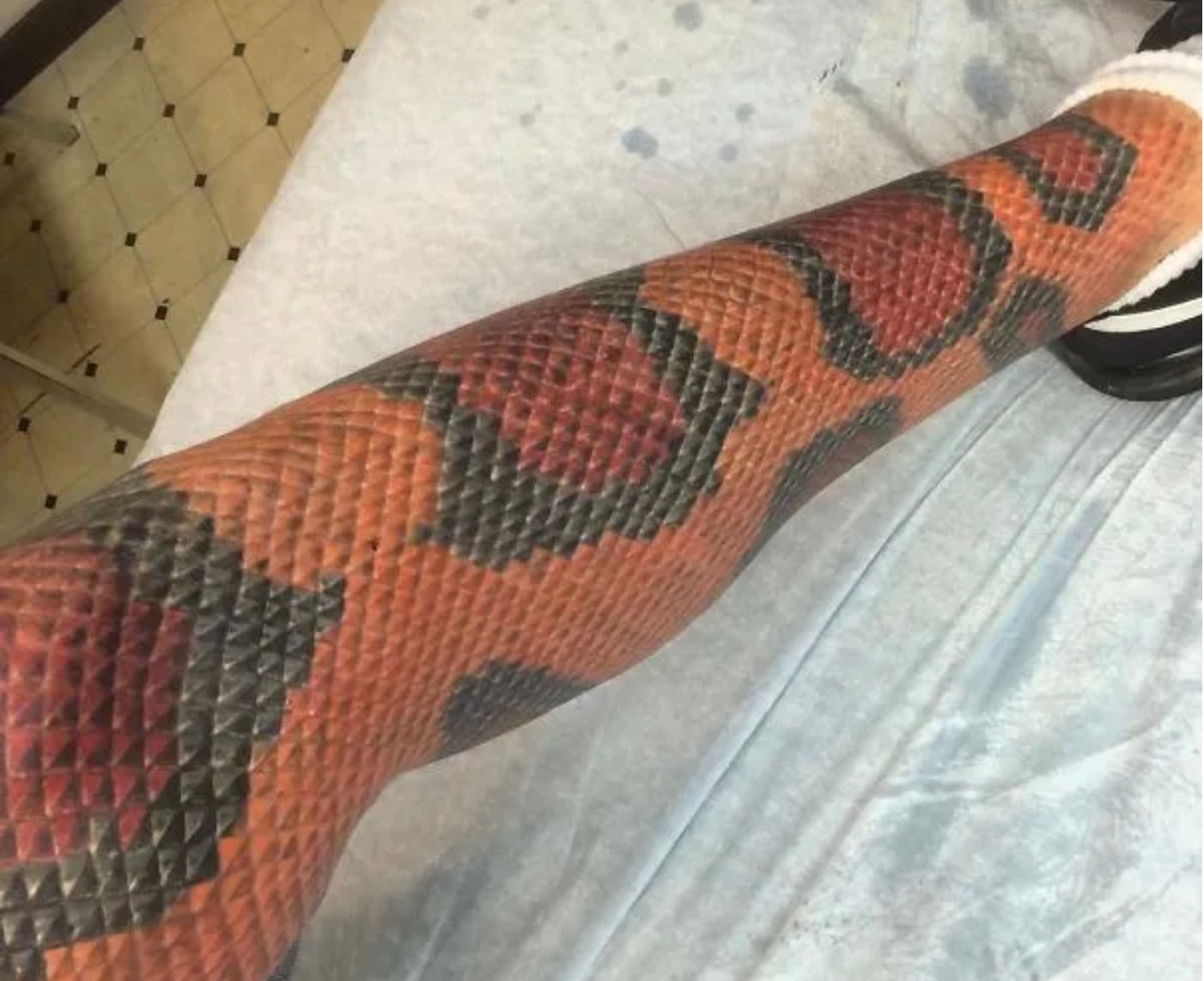 A leg tattooed to look like a snake’s skin, with detailed red, black, and orange scales, resting on a medical sheet. The realistic design mimics a snake wrapped around the leg.