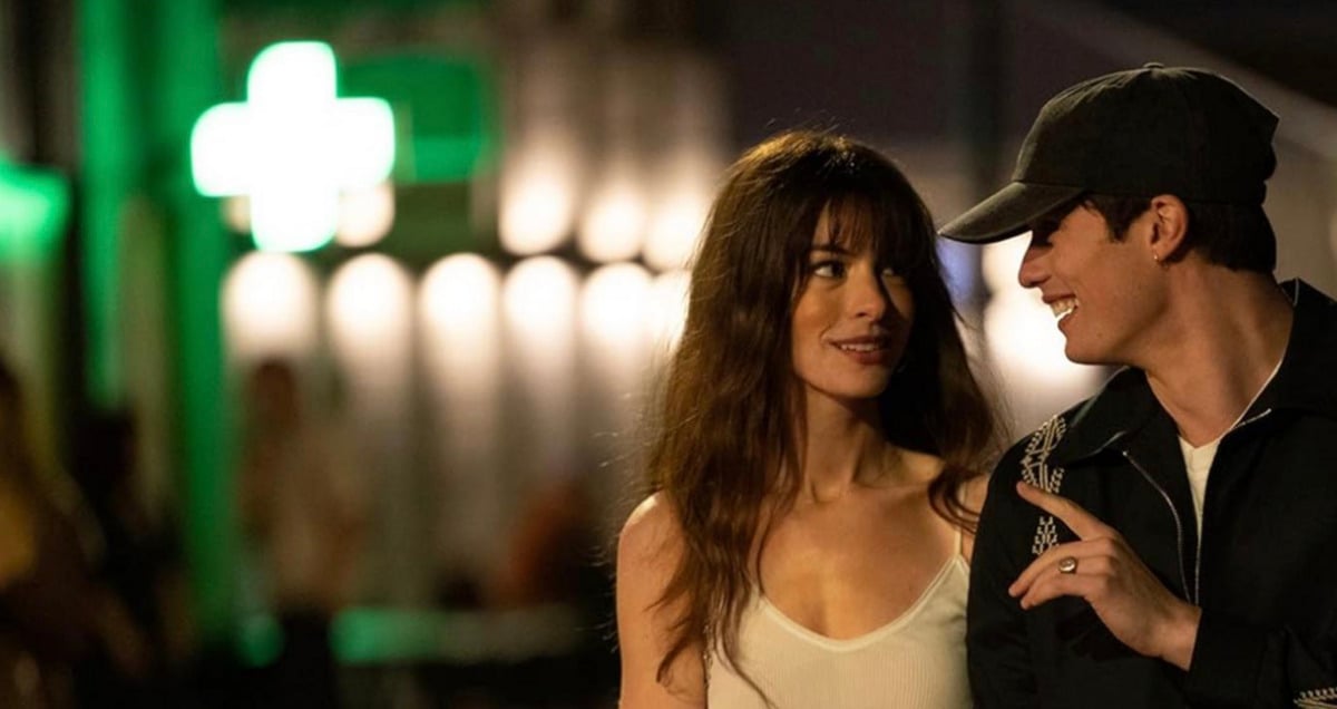 A smiling man in a dark jacket and baseball cap talks to a woman in a white tank top at night. They walk together outside under green and white lights, with a blurred neon cross sign in the background.