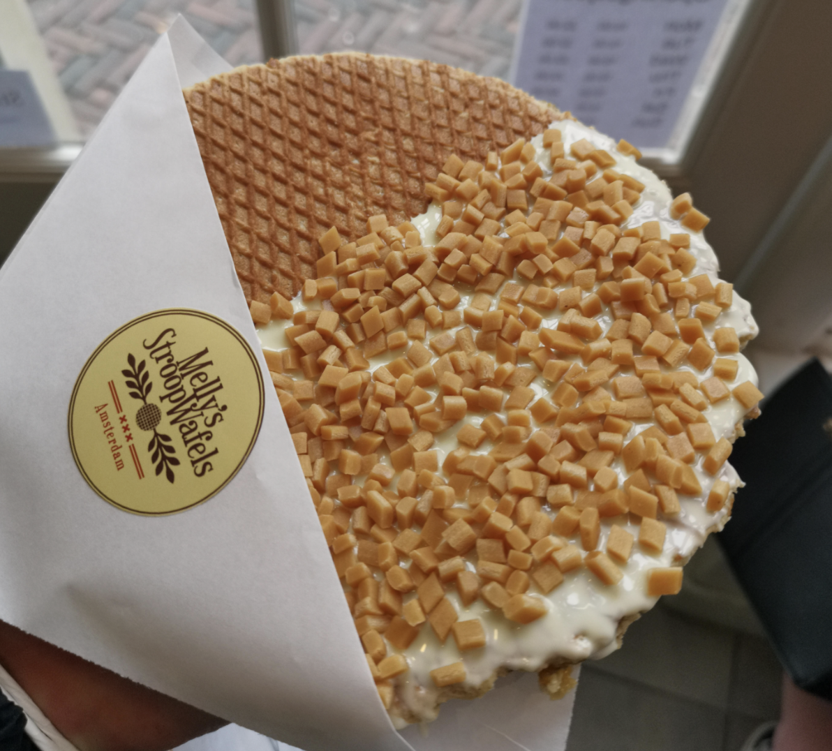 A large stroopwafel in a paper sleeve, half covered with white chocolate and small caramel cubes. The sleeve has a round sticker labeled "Melly’s Stroopwafels." A window with a blurred menu is in the background.